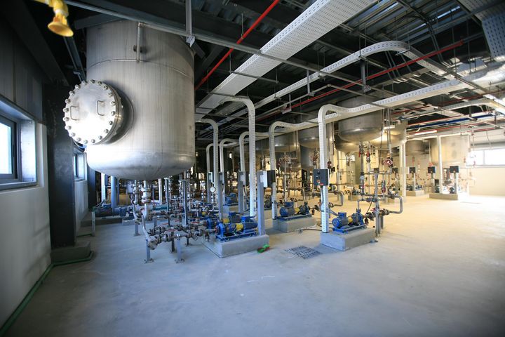 Production hall with a large cylindrical stainless-steel vessel on the left and several blue pumps on the floor. Pipes and cable trays run overhead.
